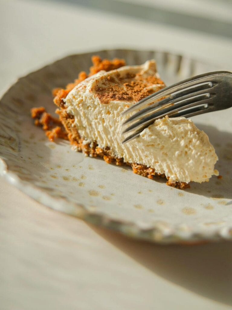 A slice of creamy no-bake pumpkin cheesecake with a graham cracker crust is placed on a light, textured plate. A fork is gently cutting into the cheesecake, and soft sunlight casts shadows on the table.